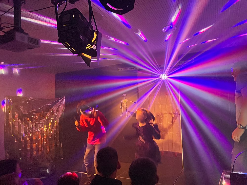 Eine Schülerin und ein Schüler tanzen auf der Bühne. Im Raum scheint eine blau-rot-gelb kreisende Beleuchtung. 