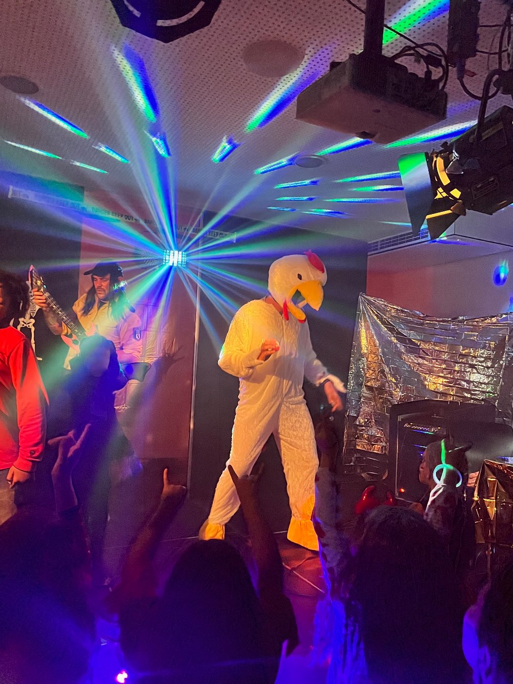 Eine als Huhn verkleidete Person tanzt auf der Bühne. Im Hintergrund sieht man blau-grün rotierende Lichter.