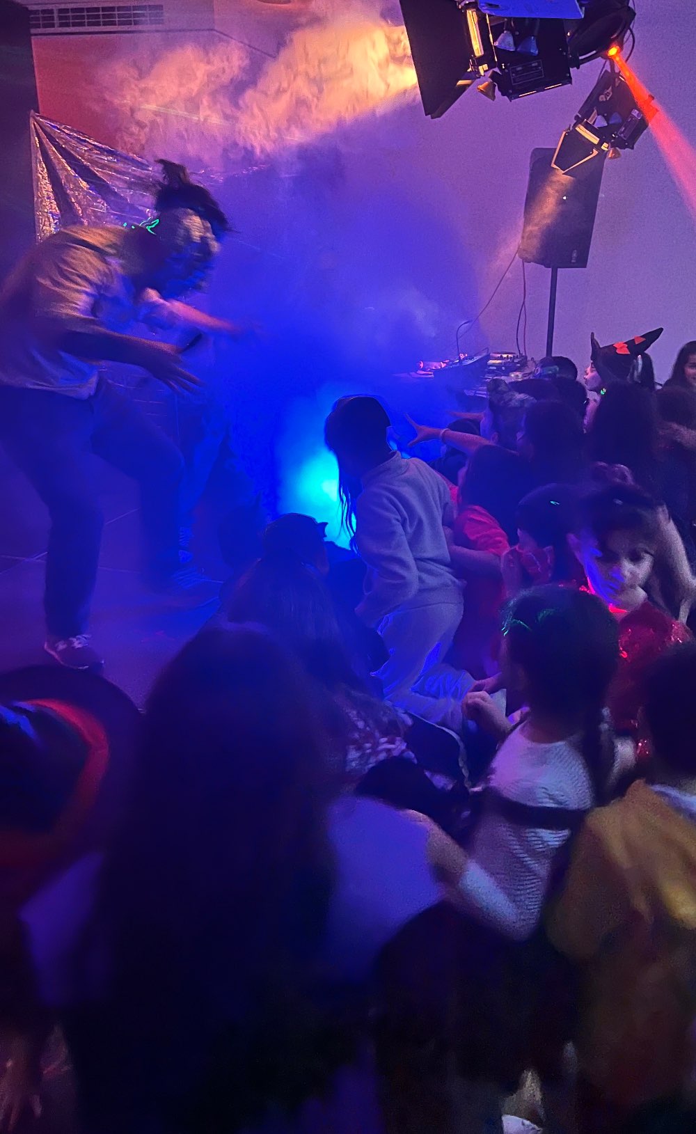 Eine als Monster verkleidete Person tanzt auf der Bühne. Vor der Bühne springen tanzend Kinder. Der Raum ist blau-pink-orange beleuchtet. 